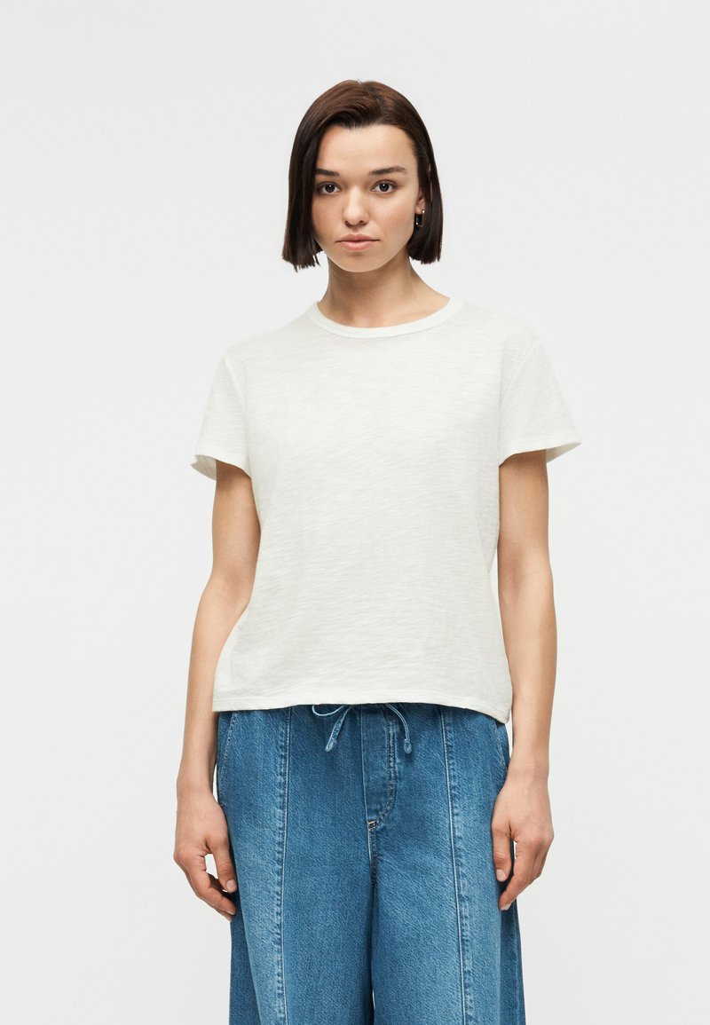 Femme aux cheveux courts bruns portant un t-shirt blanc uni à manches courtes et un pantalon en jean bleu ample sur un fond gris clair.