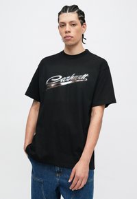 Czarny bawełniany t-shirt z metalicznym, srebrnym logo "Carhartt", krótkimi rękawami i luźnym krojem. Noszony z dżinsami.