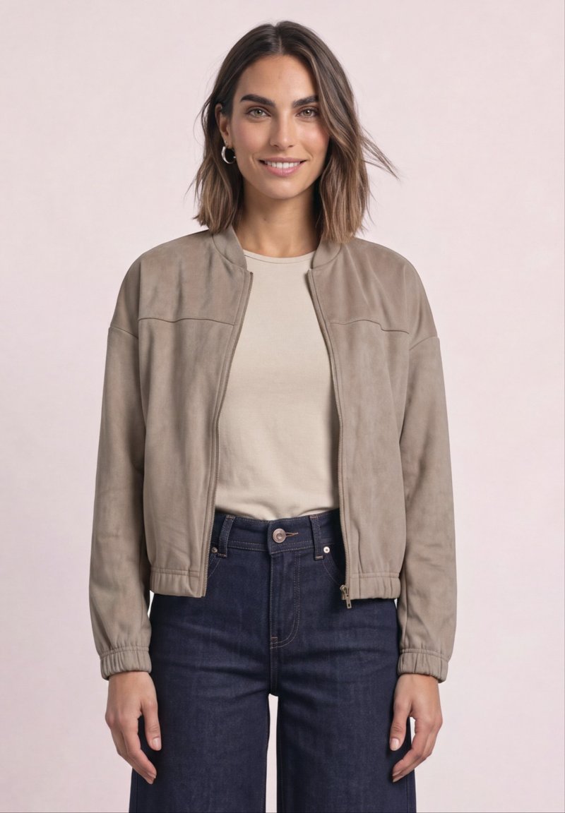 Femme aux cheveux bruns mi-longs portant des boucles d'oreilles en argent en forme d'anneaux, une veste beige zippée, une chemise crème et un jean bleu foncé sur un fond clair.