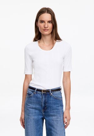 Femme aux longs cheveux bruns portant une chemise blanche à manches courtes rentrée dans un jean bleu taille haute avec une ceinture noire, debout face à l’avant.