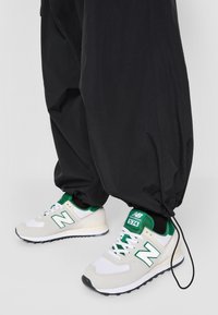 New Balance 574 sneakers with a white mesh and suede upper, green accents, and black outsole, paired with loose black tapered pants.