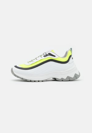 Zapatillas deportivas gruesas blancas con acentos de malla amarilla y negra, suela de goma gris texturizada, cordones reflectantes y una silueta distintiva en forma de ola.