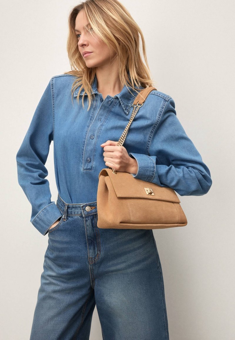 Brun ruskinds-taske med fold-over klap og gulddetaljer, kombineret med en blå denimskjorte og højtaljede jeans.