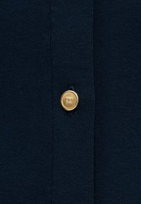 Ύφασμα πλεκτό σε ναυτικό μπλε με υφασμένο μοτίβο, που διαθέτει στρογγυλό, χρυσό κουμπί με διακριτικό σχέδιο. Κοντινή λήψη με ορατές λεπτομέρειες ραφής.