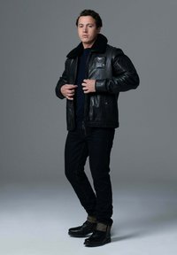 Veste en cuir noire avec col en shearling, fermeture éclair à l'avant et poches latérales. Portée sur une chemise noire avec un jean noir et des bottes noires.