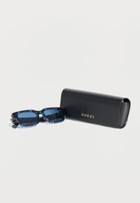 Gafas de sol rectangulares con estampado azul junto a un estuche de cuero negro con el logo dorado de Gucci sobre un fondo blanco.