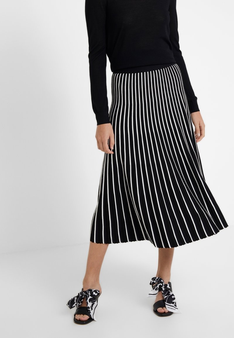 Femme porte un haut noir à manches longues et une jupe midi rayée verticale en noir et blanc avec des talons ouverts noirs attachés avec des foulards à motifs noir et blanc.