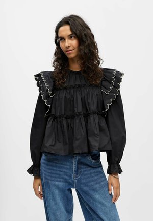 Blusa nera con maniche arricciate a bordi scallop, livelli arricciati e maniche lunghe, abbinata a jeans blu a vita alta.