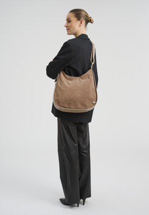 Žena s vezanom kosom nosi crni sako i hlače, nosi veliku smeđu torbu od brušene kože na ramenu, na jednostavnoj pozadini.
