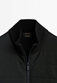 Veste matelassée noire avec col montant côtelé et fermeture éclair frontale, affichant une étiquette de marque et une texture de tissu lisse.