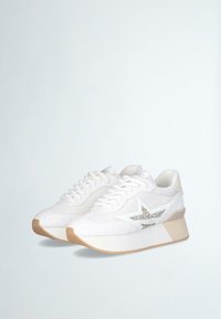 Scarpe da ginnastica bianche con una tomaia texture, caratterizzate da un accento a forma di stella argentata, design con lacci e suola in gomma leggera con un leggero tacco.