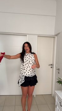 Weißes Top mit schwarzen Polka-Dots, präsentiert mit einem drapierenden Design. Kombiniert mit schwarzen Rüschen-Shorts. Ein rotes Accessoire wird in der linken Hand gehalten.