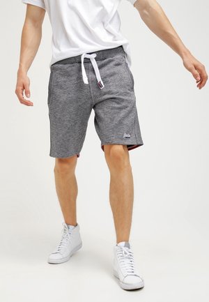 Mężczyzna ubrany w szare szorty z sznurkiem, białe sneakersy za kostkę i białą koszulkę, stojący z lekko rozszerzonymi ramionami.