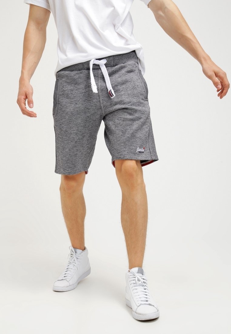 Homme portant un short gris à cordon, des baskets montantes blanches et un t-shirt blanc, debout avec les bras légèrement étendus.