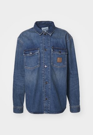 Chemise en denim à manches longues, de couleur bleu moyen, avec deux poches poitrine à boutons-pression et une patch en cuir marron sur la poche gauche.