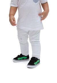 Sneaker per bambini neri e verdi con strap in Velcro, caratterizzati da un design a goccia verde. Jeans skinny bianchi strappati e una maglietta bianca a maniche corte.