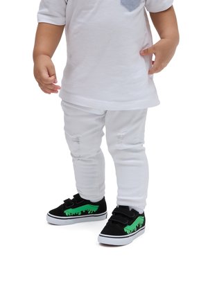 Sorte og grønne sneakers til tumlinge med velcro-stropper, præget af et grønt dryp-design. Hvide slidte skinny jeans og en hvid t-shirt med korte ærmer.