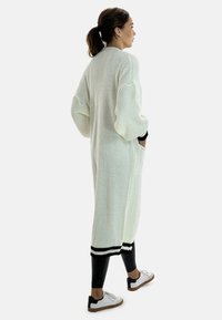 Cardigan lungo di colore crema, lavorato a maglia, con una vestibilità ampia, tasche laterali e dettagli a coste in bianco e nero all'orlo, abbinato a leggings neri e sneakers.