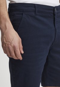 Námornícko modré šortky vyrobené z mäkkej textílie. Obsahujú predné vrecká, zapínanie na gombík a strih, ktorý sa dobre prispôsobí s hladkou textúrou.