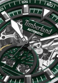 Cadran vert avec des éléments en argent, présentant un mouvement mécanique complexe et un design de logo en forme d'arbre. Marquages clairs et style moderne.