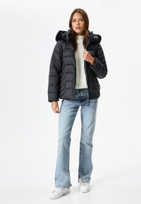 Schwarze Daunenjacke mit Kapuze und Kunstfellbesatz, mit gestepptem Design und Reißverschluss, getragen über einem weißen Strickoberteil und hellen Jeans.