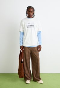 White t-shirt with a graphic print, layered over a light blue long-sleeve shirt, paired with brown checkered wide-leg trousers and a brown bag.