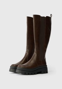 Bruna knähöga stövlar i slätt läder med elastiska sidopaneler, en kraftig svart platåsål och en dragflik bak.