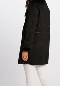 Sort suede frakke med faux fur krave og manchetter. Har lodrette sømme og sidelommer. Bæres over lyse bukser.