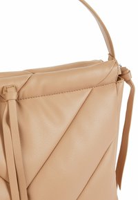 Sac à main en cuir matelassé beige avec surpiqûres diagonales, une petite anse, et des liens décoratifs en cuir sur fond blanc.