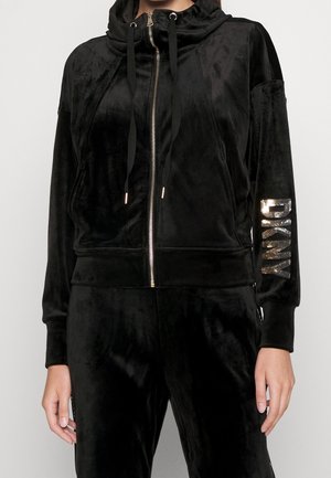 Person trägt schwarzen Velours-Zip-Hoodie mit Kapuze und langen schwarzen Kordeln, mit goldenen "DKNY"-Buchstaben auf dem linken Ärmel.