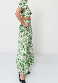 Crop top floral vert avec des manches bouffantes associé à une longue jupe à volants. Les deux présentent divers motifs botaniques verts. Des talons noirs complètent le look.