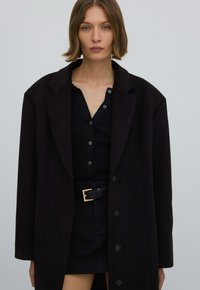 Zwarte wollen jas met een gestructureerd ontwerp, grote knopen en een notched revers, gedragen over een zwart geribd topje en rok met een ceintuur in de taille.