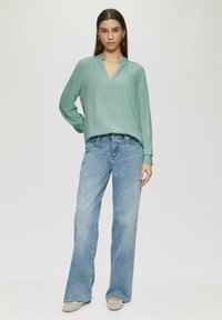 Hellgrüne Bluse mit langen Ä sleeves und V-Ausschnitt, kombiniert mit hellblauen, weit geschnittenen Jeans. Das Outfit wird von beigen Espadrilles ergänzt.