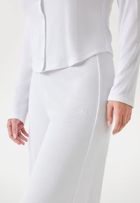 White ribbed long-sleeve top with buttons and matching pants featuring a subtle logo. Material appears soft and stretchy with a textured finish.