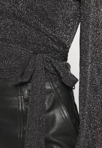 Svart topp med en glänsande texturerad tyg, som har långa ärmar och en knytning i midjan. Kombinerad med släta svarta läderbyxor.