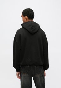 Άνδρας με κοντά, σγουρά μαλλιά, που φοράει μαύρο φούτερ με κουκούλα και σκούρα τζιν, στέκεται με την πλάτη προς το μέρος μας μπροστά σε ένα απλό ανοιχτό γκρι φόντο.