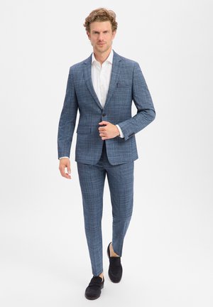 Man loopt vooruit, draagt een blauw geruit pak, wit overhemd en zwarte loafers op een witte achtergrond.