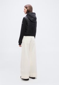 Γυναίκα που κοιτάζει προς τα αριστερά, φοράει μαύρο φούτερ με κουκούλα και μακριά μανίκια καθώς και λευκό φαρδύ παντελόνι ψηλής μέσης, στέκεται πάνω σε απλό ανοιχτό φόντο.