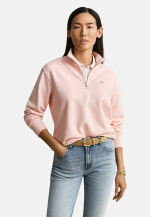 Vrouw met lang donker haar, gekleed in een lichtroze trui met een kwart rits over een wit shirt, blauwe spijkerbroek en een beige geweven riem.