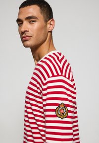 Rood-witte gestreepte longsleeve shirt met een ronde halslijn en geborduurd logopatch op de linkerarm. Zachte katoenmix stof.