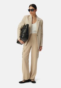 Beige gestreepte blazer over een witte tanktop, gecombineerd met beige cargobroek en zwarte platte schoenen. Het model houdt een zwarte puffer tas vast en draagt een zonnebril.