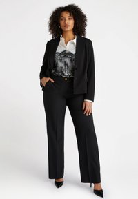 Svart skräddarsydd kavaj med en vit skjorta med svart spetsmönster, i kombination med svarta vida byxor och svarta högklackade skor.