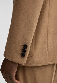 Ärmel eines braunen Wollblazers mit drei schwarzen Knöpfen, glatter Textur und taillierter Passform mit sichtbaren Nähten.
