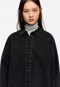 Zwarte denimshirt met zilveren studs langs de schouders, voorzien van een puntige kraag en een knoopsluiting aan de voorkant. Grijze turtleneck eronder.