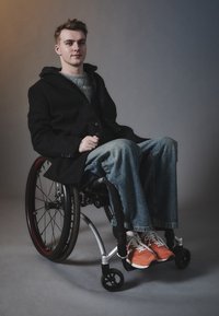 Schwarzer Mantel über einem grauen Pullover, blaue weite Jeans und orangefarbene Sportschuhe an einer Person in einem modernen Rollstuhl mit rot akzentuierten Rädern.