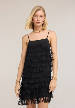 Blondynka w czarnej, bezrękawnikowej sukience z frędzlami i cienkimi ramiączkami, złotym naszyjnikiem, kolczykami-kołami i wieloma złotymi bransoletkami.