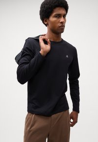 Tricou lung cu mâneci negre, în design ajustat, având un mic logo alb pe piept, fabricat dintr-un material de bumbac neted.