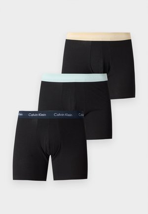 Model mężczyzna stoi boso na białym tle, nosząc czarne bokserki Calvin Klein, trzymając prawą rękę lewą dłonią.