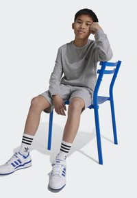 Grauer Langarm-Sweatshirt und Shorts, sitzend auf einem blauen Stuhl. Weiße Sneakers mit blauen Akzenten und gestreiften Socken. Einfaches Design und neutrale Farben.