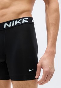 Ανδρικός κορμός και χέρι που φοράνε μαύρα συμπιεστικά σόρτς Nike με εμφανές λογότυπο στη μέση και το πόδι, σε απλό φόντο.